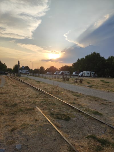The image depicts a campsite in Béjar, Spain, with several recreational vehicles parked on a gravel lot. In the foreground, disused railway tracks run through dry, grassy terrain. The sun is setting, casting a warm glow and creating dramatic light rays through the clouds in the sky. Trees line the background, forming a dense green backdrop against the sunset. The scene appears peaceful and serene, with no people visible. The overall impression is one of a quiet, natural setting at the end of the day.