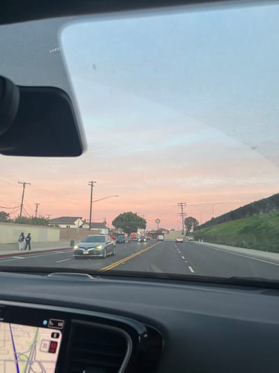 From a car's interior, a road stretches ahead under a soft pink sky. Two lanes are visible, with vehicles including a silver sedan and a truck in the distance. Utility poles line the roadside, with buildings and a tree partially obscured. A grassy slope rises on the right, and the car's dashboard and navigation screen frame the view. People walk on a sidewalk beside the road.