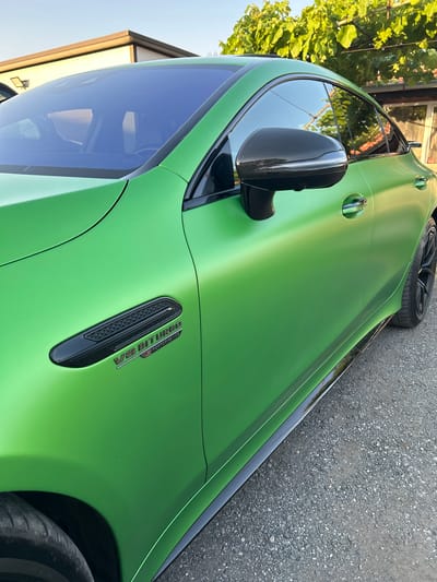 This is a close-up, angled shot of a matte green Mercedes-AMG GT 63 S E Performance parked on a gravel surface in Slanchevo, Bulgaria. The vehicle is the primary subject, with a focus on its side profile, including the front fender, door, and side mirror. The car features a carbon fiber side mirror and a prominent "V8 BITURBO E PERFORMANCE" badge on the fender. The setting appears to be outdoors during the daytime, with a hint of clear sky and some greenery from a vine in the upper left corner. The gravel ground suggests an outdoor parking area or road. The lighting indicates it is likely late afternoon or early evening, with sunlight casting a soft glow on the car's surface. There are no people visible in the frame.