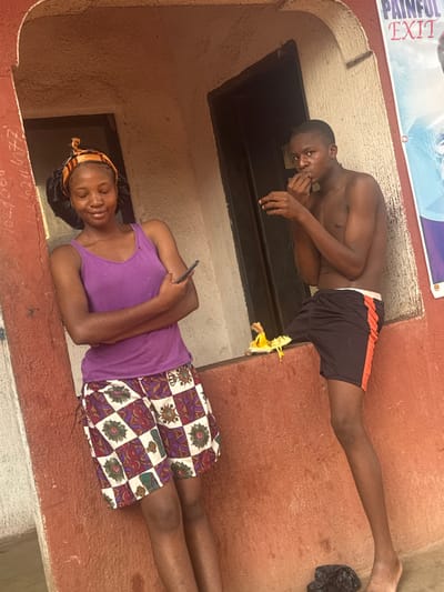 Two young people are positioned on a ledge in front of a dark rectangular opening in a beige wall. The person on top is a shirtless young man with short hair, wearing black shorts with an orange stripe. He is looking at the camera. The person below is a young woman in a purple tank top and patterned shorts, holding a smartphone and looking down, smiling gently. A yellow flower sits near the top of the ledge. A sign reading "PAINFUL EXIT" is visible in the background.