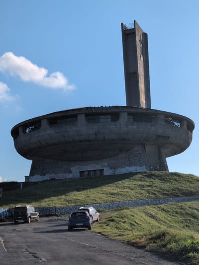This is a media file depicting the Buzludzha Monument, a former communist monument in Bulgaria. The monument, a large, Brutalist concrete structure resembling a UFO or a spaceship, is situated on a grassy hillside. Its iconic disc-shaped main hall and a tall tower are visible against a bright blue sky with a few white clouds. The monument appears abandoned and weathered, with some signs of decay.
Below the monument, a paved road runs alongside the hillside, which is covered in lush green grass and a stone retaining wall. Several cars are parked on the roadside, suggesting that visitors are present. The scene is bathed in bright sunlight, indicating a clear day.
The original image is oriented vertically, with the monument dominating the left side and the road and hillside on the right. The visible text on the monument is in Cyrillic script, likely containing slogans or historical inscriptions related to the communist era. The overall impression is one of a monumental and somewhat eerie relic of the past set against a natural landscape.