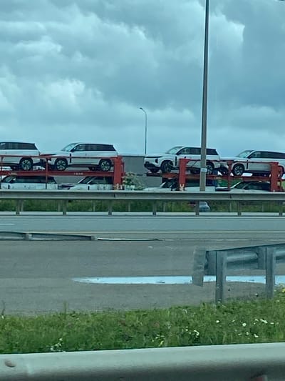 The image shows a car carrier trailer loaded with several white SUVs on a highway under a cloudy sky. The cars appear to be new and are stacked on two levels. In the foreground, there is a road with a white dividing line and some green grass and weeds. There is also a reflective barrier or guardrail. The car carrier is positioned on the right side of the road. The location is identified as Kliuchiki, Russia.