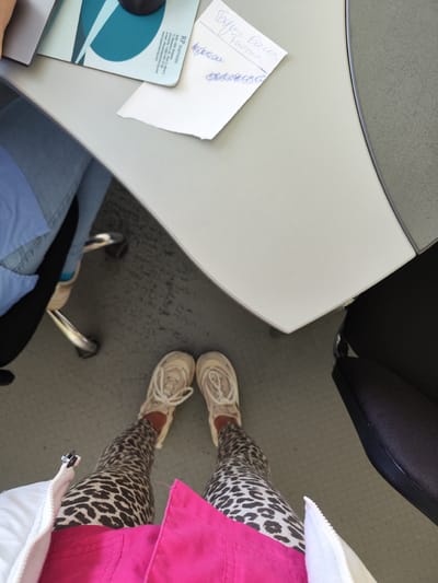 A first-person perspective shot depicts someone standing in what appears to be an office setting. The person is wearing leopard-print leggings, cream-colored sneakers with white laces, a white jacket, and a bright pink garment, likely a shirt or jacket underneath. The setting includes a gray desk with a mouse pad and a piece of paper with "Борис Баш Tuomar" written on it, and what looks like a business card for "R.P. Partners". The floor is covered with a gray patterned material. Another person is sitting near the desk. Based on the business card, the location is likely Sofia, Bulgaria, not Aprilovo.