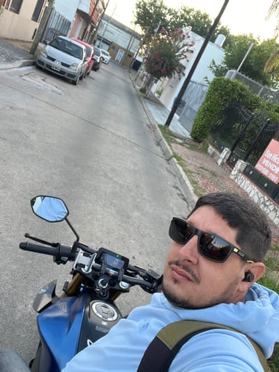 The image depicts a male subject seated on a stationary blue and black Honda motorcycle on a paved residential street in Bernal Este, Argentina. The subject, estimated to be in his 20s or 30s, is positioned in the foreground, looking towards the camera. He wears dark rectangular sunglasses with "GLOX" branding, a light blue hooded sweatshirt, and a dark green backpack. A black wireless earbud is visible in his left ear. The motorcycle's digital dashboard displays a speed of "0," and a mobile phone holder is attached to the handlebars.
The street, sloping gently, is flanked by concrete sidewalks. On the right, a residential property features a black metal fence partially obscured by a green hedge. A red and white real estate sign is visible on this property, displaying "San Nico VENDE" and contact numbers "4207-0350" and "5180." In the background, along the left side of the street, multiple parked vehicles include a silver Renault Clio and a red hatchback. Various residential buildings line both sides of the street, interspersed with green trees and flowering bushes. A utility pole stands on the right sidewalk. The scene is illuminated by daylight, suggesting late afternoon or early evening.