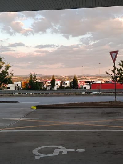 The image captures a parking area for semi-trailer trucks, likely a rest stop or depot, under a soft, cloudy sky at dusk. Several white and red trucks are parked in a row, some with trailers, indicating a commercial trucking operation. The foreground shows a paved surface with yellow and white lines, designating parking spaces, and a white symbol for disabled parking is visible on the ground. A red and white triangular yield sign is present on the right side of the image, along with a tree. In the background, rolling hills and distant structures suggest a rural or semi-rural setting. The overall impression is one of a functional transportation hub at the end of the day. The specific location in Osek nad Bečvou, Czechia, is not visually evident from the image alone but would be external knowledge.