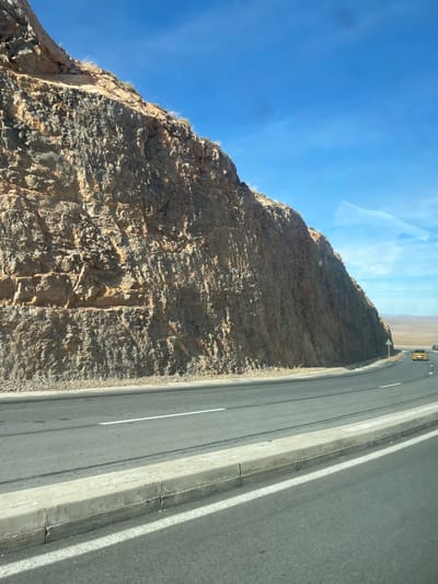 The image displays a curved asphalt road situated alongside a prominent, rugged rock formation near Amersid, Morocco, under a clear blue sky. The rock face, occupying the left and central frame, exhibits multi-toned brown and dark grey strata with visible geological textures and minimal dry vegetation along its upper edges. The two-lane road features a dashed white centerline and a solid white shoulder line, curving sharply to the right and receding into the distance. A segmented concrete curb runs parallel to the road in the immediate right foreground. A single yellow vehicle is observed driving away from the viewer on the distant curve of the road. A white triangular road sign is positioned on the roadside beyond the yellow vehicle. The distant background reveals an arid landscape with rolling hills. No individuals are present in the scene. Reflections on the upper right side suggest the photograph was taken from inside a vehicle.