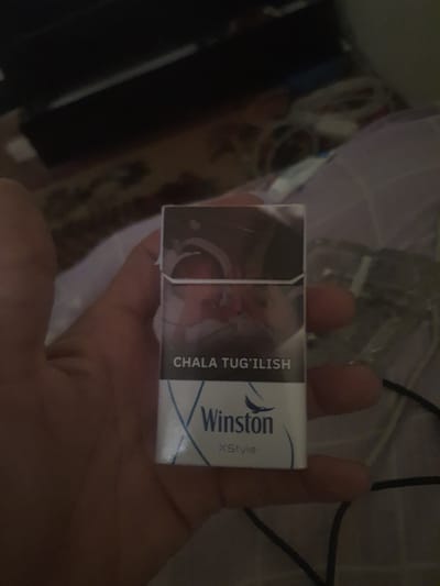 A hand holds a pack of Winston cigarettes. The pack is mostly white with blue accents and text. On the side of the pack, vertical text reads "CHALA TUG'ILISH". Below the Winston logo, the text "XStyle" is visible. The background is blurry and dark, suggesting an indoor setting, possibly a dimly lit room. The lighting is not strong, and there are shadows present. The image appears to be taken at close range, focusing on the cigarette pack and the hand holding it.