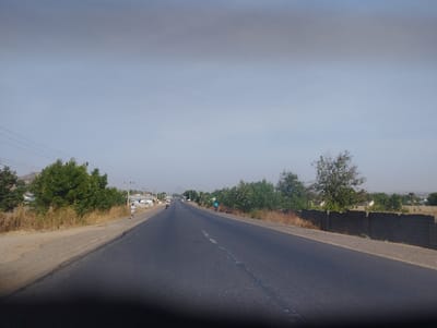 The image shows a straight, paved road with a single lane in each direction, located in Kaltungo, Nigeria, at the coordinates 9.825 latitude, 11.318 longitude. The road runs straight through a rural area with a low wall on the right side of the road. The wall is made of grey concrete blocks. On the left side of the road are a few trees and bushes.  There are two people walking in the distance on the right side of the road. One person is walking toward the camera. The other person is walking away from the camera. It appears the sky is a light grey color and the time of day is midday.  The road appears to be empty with the exception of the two pedestrians.
