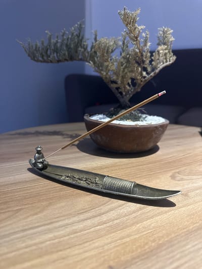 The image depicts a still life arrangement on a wooden table. In the foreground, a metallic incense holder, shaped like a narrow boat with embossed decorative elements, holds an unlit incense stick. A small, metallic figurine of a seated person wearing a hat is affixed to the left end of the incense holder. The incense stick extends diagonally across the frame, leading towards a bonsai tree in a brown ceramic pot in the midground. The bonsai tree appears dry and sparse, with white material, possibly sand or small pebbles, in its pot. In the background, a dark, out-of-focus sofa is visible, and the wall behind it is a muted blue. The scene is illuminated by ambient light, casting subtle shadows. There is no indication of the city of Zibatá, Mexico.