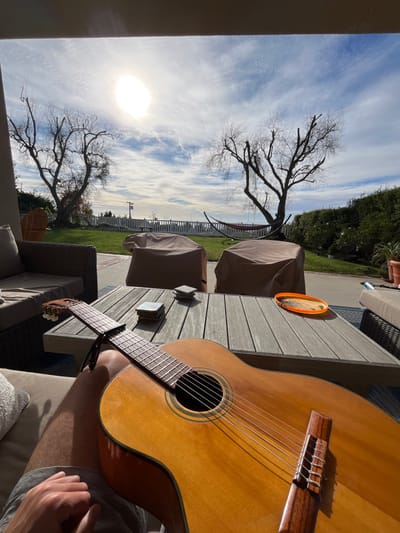 An outdoor setting with a guitar as the focal point, held by a person seated on a patio. A wooden table holds a frisbee and coasters. The background features a yard with two trees, a white picket fence, and furniture covered with protective cloth. The sky shows a bright sun and thin clouds.