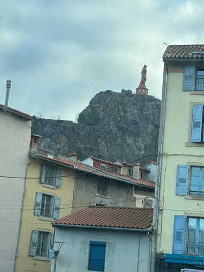 The image depicts an urban scene in Le Puy-en-Velay, France, dominated by the Rocher Corneille with the Statue of Notre-Dame de France under an overcast sky. In the background, the dark, rugged volcanic peak of Rocher Corneille rises prominently. Perched atop this peak is the reddish-brown statue of Notre-Dame de France, depicting the Virgin Mary and Child, resting on a multi-tiered circular pedestal. Sparse, bare trees are visible on the slopes of the rock, and a winding path can be discerned. The sky is uniformly light gray and cloudy.
In the foreground and midground, multiple residential or mixed-use buildings line the lower parts of the city. These structures feature tiled roofs, primarily in shades of terra cotta orange. Building facades are painted in various hues, including pale yellow, light gray, and off-white. Many windows are fitted with light blue louvered shutters, some open, some closed. A tall, slender metal chimney pipe extends from a roof on the far left. A blue awning with partially visible white lettering, "TAJMAHA," indicative of a commercial establishment, is present at the bottom right. Overhead wires traverse the scene, extending across the buildings. No individuals are visible, and no discernible actions or interactions are depicted.