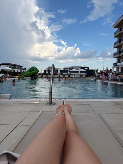 A first-person perspective shot captures a relaxed scene by a swimming pool. In the foreground, a person's legs and feet, adorned with pink nail polish, are stretched out on a light-colored lounge chair. The pool, with its blue tiled edge and clear water, is to the left. A metal railing extends into the pool.
Beyond the pool, the background reveals a resort-like setting. People are seen swimming and relaxing by the water. A bright green inflatable slide shaped like a dragon is a prominent feature in the pool. Buildings with modern architecture and white umbrellas dot the landscape. The sky above is a vibrant blue with scattered white clouds, suggesting a pleasant day, possibly late afternoon given the lighting.
The overall mood is one of leisure and enjoyment, characteristic of a vacation or a leisurely day at a hotel pool. The location is identified as Hisarya, Bulgaria. Visible text includes "SENATE" on a white sign.