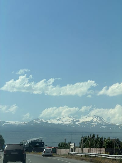 In the city of Proshyan, Armenia, under a clear blue sky with scattered white clouds, a highway passes by a landscape dominated by snow-capped mountains in the distance. A black Jeep is driving on the highway, with another car following behind. To the right of the highway, a modern building with a dark, angular roof is visible, along with a fence and various trees. The scene captures a moment of travel through a scenic Armenian location.