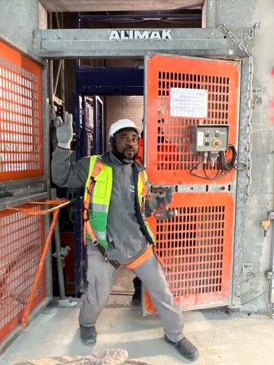 The image displays a male construction worker posing inside a building under construction in Aulnay-sous-Bois, France. He is positioned in front of an open Alimak industrial hoist system, identified by the "ALIMAK" branding above the entry. The man, with a beard and wearing a white hard hat, ear protection, a grey long-sleeved top, a high-visibility yellow and green safety vest with an attached harness, grey work pants, and dark work boots, is looking directly at the camera with a slightly surprised expression. He wears grey gloves and holds a cordless drill in his right hand. His left arm is raised, with two fingers extended in a peace or victory gesture. The hoist features orange grated safety doors, with one partially open on the left and another fully open on the right, revealing the interior shaft. A control panel with a red emergency stop button and two black buttons is mounted on the inside of the right orange door. A white sign on this door displays French text instructing to contact security for door opening. The floor is bare concrete with minor debris, and concrete structural elements are visible in the background.