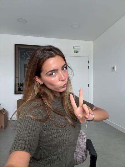 A young woman is captured in a selfie, with her head tilted to the side and a slight pucker on her lips, looking directly at the camera. She holds up two fingers in a peace sign with her left hand, which displays a silver-colored bracelet. Her long, brown hair with lighter highlights frames her face and falls over her shoulders. She is wearing a textured, dark green knit sweater. A small, silver-toned cross earring is visible in her right ear.
The setting appears to be a modern indoor space, possibly an office or a bright room in Torres Vedras, Portugal. The walls and ceiling are white, providing a clean backdrop. In the background, a white door is visible, above which a green emergency exit sign with an arrow pointing left and a human figure can be seen. A white square device, possibly a smoke detector, is mounted on the ceiling, along with a circular, recessed light fixture. A framed artwork featuring a dark geometric pattern hangs on the wall behind her left shoulder. Part of a grey office chair is visible to her right, and a brown cardboard box with faint markings, including "II" in a white square, is present in the lower right corner. The flooring appears to be a grey, possibly concrete or tiled surface. The lighting is bright and even, consistent with an interior space during the day. The text "happe" is indistinctly visible on the silver bracelet.