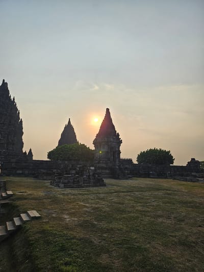 The image depicts the Prambanan Temple complex in Tlogo, Indonesia, at sunset. The sky is a pale yellow with the sun visible as a bright orb, partially obscured by clouds. Several tiered stone structures, characteristic of ancient Indonesian temples, dominate the scene. The largest structures are tall, pyramid-like towers with intricate stone carvings. In the foreground, a grassy field stretches out, with some scattered stone ruins and a set of stone stairs leading up a slight incline on the left. Lush green trees are visible between the temple structures. The overall lighting suggests the golden hour of sunset, casting a warm glow on the scene and creating long shadows. There are no people visible in the image.