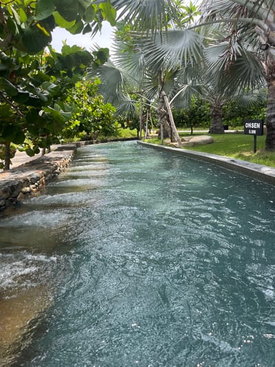 The image depicts a man-made water channel flowing through a tropical landscape. The channel is lined with stone and concrete, with water flowing gently, creating ripples and small waves. On the left side of the channel, there is a stone wall with lush green foliage overhanging. On the right side, the channel is bordered by a well-manicured lawn, with palm trees and other tropical vegetation visible in the background. A black sign with white lettering reads "ONSEN 0.9M", indicating a depth of 0.9 meters and that this is likely an onsen, or hot spring. The sky is overcast, suggesting it might be a humid or partly cloudy day. The overall scene conveys a sense of tranquility and relaxation, typical of a resort or spa environment in a warm climate. The location is identified as Cam Lam, Vietnam. There are no people visible in the image.