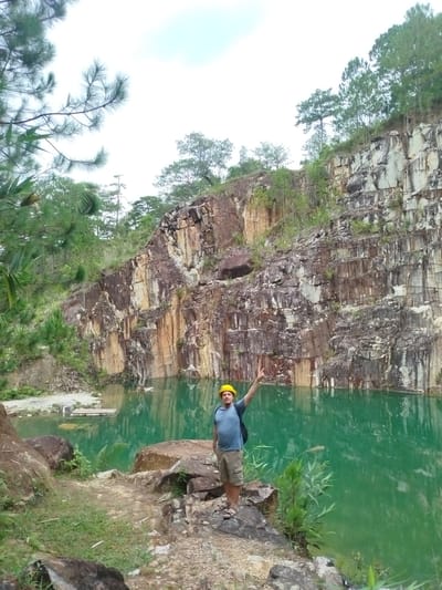 The image depicts a man wearing a yellow hard hat, a blue t-shirt, and khaki shorts, standing on a rocky outcrop. He is raising his left arm, making a peace sign with his fingers. The man is positioned to the left of the frame, with a vibrant turquoise body of water stretching out before him. In the background, a towering cliff face, characteristic of a quarry, rises steeply. The cliff exhibits distinct layers of rock, with patches of vegetation growing from its crevices. The water reflects the sky and the surrounding cliffs, creating a striking visual contrast. The overall environment suggests an outdoor, natural setting, likely a former quarry repurposed as a scenic location in Lán Tranh, Vietnam.