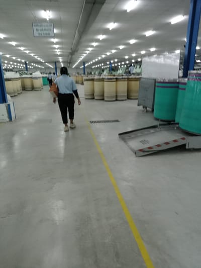 This image depicts the interior of a large industrial facility, likely a textile or yarn production factory, located in Nglundo, Indonesia. The scene is dominated by a wide, well-lit aisle with a light gray concrete floor, marked by a bright yellow line running down its center.

In the foreground, a person is seen from behind, walking away from the camera. They are dressed in a light blue short-sleeved shirt, dark trousers, and light-colored sneakers, carrying a light brown bag over their shoulder. Another person is partially visible further down the aisle, also appearing to walk.

On both sides of the aisle, rows of numerous large, cylindrical bobbins or spools, mostly tan or yellowish, are neatly arranged, suggesting the presence of yarn or thread. On the right side, closer to the viewer, are several taller cylindrical objects wrapped in teal plastic with purple bands, possibly larger spools or material containers. A gray industrial cart and a metal ramp with red and white striped markings, likely used for material handling, are also visible on the right.

The ceiling is brightly illuminated by numerous rectangular fluorescent lights, suggesting active operations regardless of external daylight. A white rectangular sign hangs from the ceiling in the mid-distance. The text on the sign, in Indonesian, reads: "MESIN HARUS SEGERA STOP JIKA HASIL KUALITAS TIDAK STANDARD," which translates to "MACHINE MUST STOP IMMEDIATELY IF QUALITY RESULTS ARE NOT STANDARD." This detail highlights the facility's emphasis on quality control and standard operating procedures within this manufacturing environment.