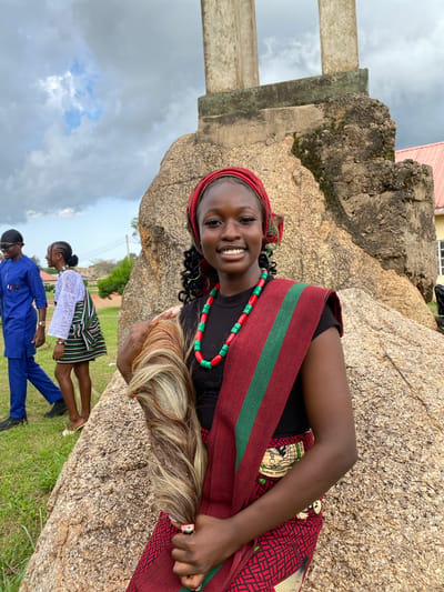 The image shows three people in an outdoor setting with large rocks and grassy ground. A young woman is prominently featured in the foreground, leaning against a rock. She has dark skin, dark curly hair, and is wearing a red headscarf. She is smiling at the camera and has a beaded necklace of red and green beads. Around her shoulders, she wears a sash or fabric with a red background and green stripes. Her right arm is visible, and she appears to be holding something light-colored, possibly hair extensions, in her hand. A ring is visible on her right index finger.
In the background, two other individuals are partially visible. One person, wearing a white top and a striped skirt, is looking down. Another person, wearing a blue outfit and sunglasses, is also visible.
The scene appears to be in a rural or natural location, with large, weathered rocks dominating the landscape. The sky is cloudy, suggesting it might be overcast or that rain could be approaching. A building with a pink roof is visible in the upper part of the image. The presence of people in traditional or festive attire and the natural setting suggest a gathering or cultural event. No visible text is present in the image. The overall mood seems cheerful, primarily conveyed by the smiling expression of the woman in the foreground.