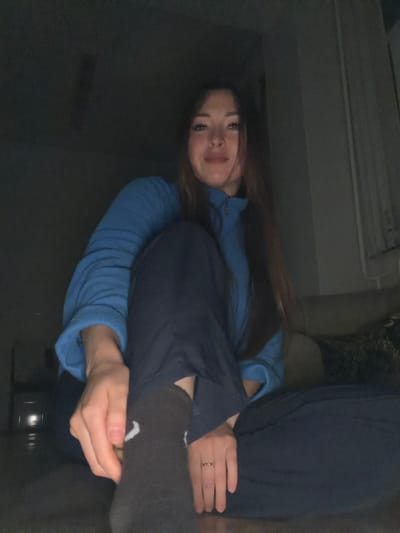 A young woman is sitting on the floor in a dimly lit room. She is wearing a blue fleece jacket and dark pants. Her legs are crossed, and she is resting her hands on her right leg. The room appears to be a living room, with a sofa visible in the background. The overall scene is casual and relaxed. There is no information in the image to suggest this location is in the city of Япрыково, Russia.