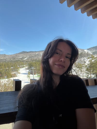 A selfie-style photograph shows a person with long dark hair, presumably female, facing the camera with a slight smile. The person is wearing a black shirt. The background reveals a mountain landscape, likely a ski resort, with snow-covered slopes and trees. In the distance, ski lifts are visible. The sky is clear and blue. A wooden structure, possibly a balcony or deck, is in the foreground. The location appears to be in a mountainous region, possibly near Озёрное, Russia.