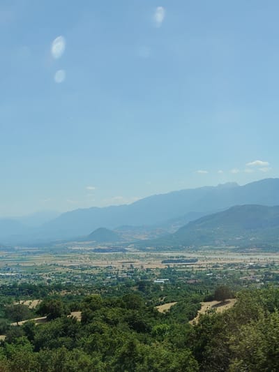 The image captures a panoramic view of the landscape surrounding Kalambaka, Greece. The foreground is dominated by lush green trees, forming a dense canopy. Beyond the trees, a valley unfolds, characterized by agricultural fields, scattered buildings, and a network of roads. In the background, a series of majestic mountains rise, their peaks fading into a hazy blue. The sky is a clear, light blue, with a few faint white clouds. Two circular, bright light artifacts are visible in the upper left portion of the sky, likely lens flares.
