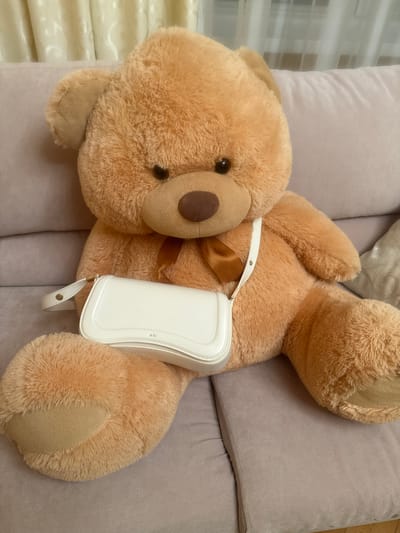 A large, plush, light brown teddy bear is seated on a pale pink couch. The bear is positioned facing forward, with its arms resting at its sides. A white, patent leather handbag with a single strap is placed in front of the bear, resting on its lap. The handbag has a gold clasp and the text "JW PEI" is subtly embossed on its front. The background consists of light beige curtains with a subtle pattern. The overall setting appears to be indoors, likely in a residential environment.