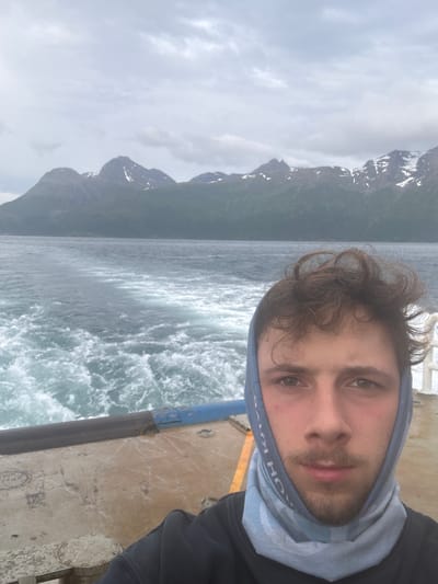 A young man with curly brown hair and light brown eyes takes a selfie on a boat. He is wearing a dark hoodie and a light blue gaiter around his neck and head. The gaiter has a pattern with text that appears to be "KICK" and "HORSE". He has a light beard and mustache. Behind him, the boat's deck is visible, made of metal with some scuff marks and a blue railing. The water behind the boat is choppy and white with foam, indicating movement. In the background, a range of mountains with green slopes and snow-capped peaks under a cloudy sky can be seen. The scene suggests a location near the city of Jektevika, Norway, likely on a ferry or other watercraft, given the wake and mountainous landscape characteristic of the region.