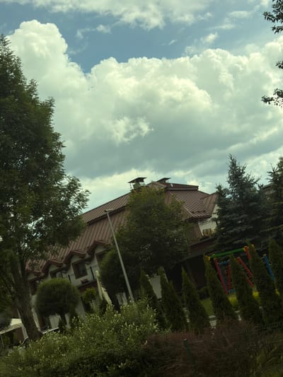 The image shows a view of a residential building in Wieliczka, Poland, with a focus on its exterior architecture and surrounding greenery. The building features a prominent brown, multi-pitched roof with dormer windows. The facade appears to be light-colored, with dark-framed windows and balconies.
Several trees and bushes surround the building, creating a natural and somewhat overgrown setting. A large tree with dense foliage stands to the left of the frame, partially obscuring the building. In the foreground, there is a variety of shrubs and smaller trees, some with white flowers. To the right of the building, a children's playground structure with colorful bars and red, yellow, and blue elements is visible. The sky above is filled with large, white, fluffy clouds against a pale blue background, suggesting a partly cloudy day. The perspective is slightly tilted, giving a dynamic feel to the scene. No people are visible in the image.