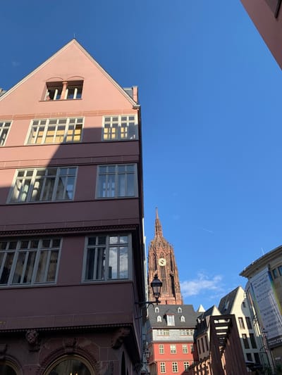 The image shows a street in Frankfurt am Main, Germany. It is a narrow street with a pink building on the left and a tall, red church spire in the background. The building is made of stone and has many windows. The windows are all closed and the building is empty. The spire of the church is topped with a clock face. The sky is blue and there are no clouds. The image is taken from street level and is focused on the building. The coordinates of the location are latitude: 50.111, longitude: 8.683. The camera lens is at a low angle. The image is taken during the day. The subject of the photo is the building. There is a street light on the left of the image. A few other buildings can be seen in the distance on the right. The image is focused on the pink building.