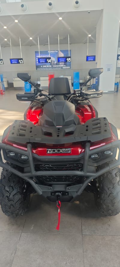 The image shows a red and black AODES brand all-terrain vehicle (ATV) parked indoors. In the background, signs indicate that the ATV is located in Bolshoye Savino Airport, Russia, with signs labeled "Пермь" (Perm) above check-in counters numbered 12 through 15. A phone number and website address are also visible.