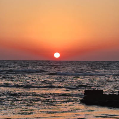 The image depicts a serene sunset over the Mediterranean Sea, likely near Mikhmoret, Israel. The sun, a bright white orb, is positioned slightly off-center in the horizon, casting a warm orange and red glow across the sky and reflecting on the water. The sea is calm, with gentle waves rolling in, their crests catching the fading sunlight. A dark, jagged rock formation is visible in the lower right corner of the frame, partially submerged by the water. The sky transitions from a pale orange at the top to a deeper red near the horizon, creating a peaceful and atmospheric scene. There are no people or specific landmarks to indicate the exact location within Mikhmoret, but the elements of the scene are characteristic of a coastal sunset.