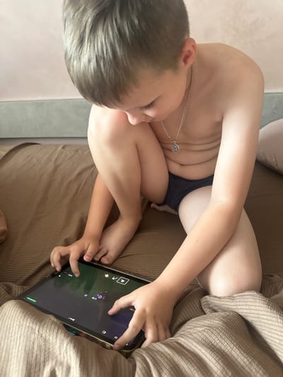 The image shows a young person playing a video game on a tablet. The person is lying down on a brown, corduroy surface, possibly a couch or bed. They are wearing dark underwear and have a silver chain necklace with a circular pendant. Their body is partially obscured by a beige, corduroy blanket. The tablet screen displays a game with purple and pink elements against a dark background, along with game controls. The person's hands are actively engaged with the tablet, indicating they are playing the game. The lighting suggests it might be daytime, but no specific time is evident. The setting appears to be indoors, in a home environment. There is no visible text in the image that provides further context.