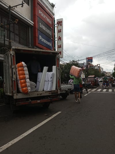 The scene depicts a commercial street in Vaatharawattai, Sri Lanka, under an overcast sky. A white box truck is parked with its rear door open, partially on the asphalt road and near a building on the left side of the frame. Inside the truck, a male individual in a grey t-shirt is visible, standing amongst goods. On the left side of the truck's cargo area, a large, rolled object, orange and white in color and marked "MULYANA," is secured. To the right, multiple bundles of white, elongated items, likely rolls of material, are stacked.
The adjacent building on the left features a prominent vertical sign structure. The top panel displays "POLYTRON" on a red background, the middle panel "SAMSUNG" on a black background, and the bottom panel "Panasonic" on a blue background. To the right of this sign, a smaller vertical sign spells "Nuhun" in red letters. An ornate, dark metal fixture is attached to the building's upper facade.
In the mid-ground, a person wearing a green long-sleeved shirt and blue shorts walks away from the truck, carrying a large, light-brown, textured, rectangular object balanced on their head. Further down the street on the right, vehicular traffic includes a red auto-rickshaw, multiple motorcycles, and a person riding a bicycle with items loaded. Overhead utility lines crisscross the sky. The road surface in the foreground shows a faded white painted line.