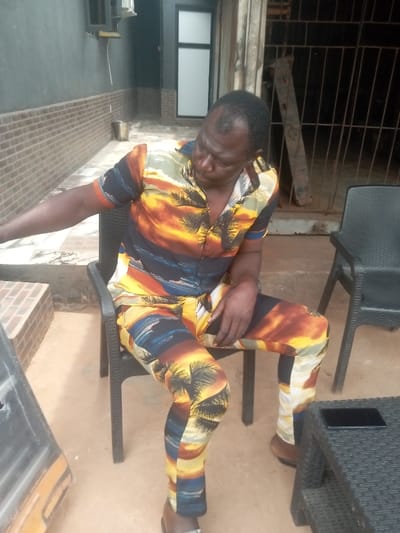 The image captures an adult male seated on a dark plastic chair in an outdoor or semi-outdoor location within Awkuzu, Nigeria. The individual, appearing middle-aged with dark skin and thinning dark hair, is dressed in a matching short-sleeved button-up shirt and trousers patterned with tropical sunset, palm tree, and ocean motifs. He is looking downwards, with his left arm extended out of frame to the left and his right hand resting on his lap. He wears dark open-toed footwear. An identical unoccupied dark plastic chair is positioned to his right, alongside a dark plastic side table on which a black smartphone rests horizontally. The environment features a building wall in the background to the left, characterized by a dark gray upper section and a brown brick-patterned lower section, with an air conditioning unit mounted and a metallic bucket visible on the ground. A white-framed door or window is also present further back. The background to the right shows a less refined area, possibly an entrance, with metal bars or a gate and leaning wooden planks. The ground consists of light brown paved sections immediately beneath the seating area, transitioning to reddish-brown earth further out.