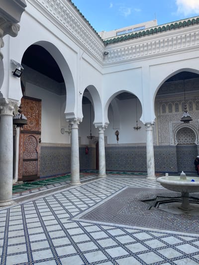This image depicts a bright, open courtyard within the Mausoleum of Moulay Idriss I in Moulay Idriss Zerhoun, Morocco. The courtyard features white-painted walls with lower sections adorned by intricate geometric zellij tilework in blue, green, and white. A series of white horseshoe arches, supported by fluted marble columns with ornate capitals, defines the perimeter of the open space, leading to internal corridors. The floor is covered in elaborate mosaic tiles, incorporating a central octagonal design around a white stone fountain with visible water, and extending into a checkered pattern of white and blue tiles.
On the left side of the frame, a tall, intricately carved wooden door is partially ajar, revealing a dark interior. Green prayer rugs are laid out on the tiled floor directly adjacent to this door. Several chandeliers, some metallic and some glass, hang from the ceilings beneath the arches. A pendulum-style wall clock is affixed to a column between two arches, and a red fire extinguisher is mounted on a wall nearby. Wall-mounted solar-powered floodlights are present on the upper white walls. Above the courtyard, a section of a green tiled rooftop is visible, with a modern building with horizontal windows partially discernible against a light blue, partly cloudy sky. Two indistinct figures, dressed in dark and lighter attire respectively, are partially visible in the far right background, near an arched opening.