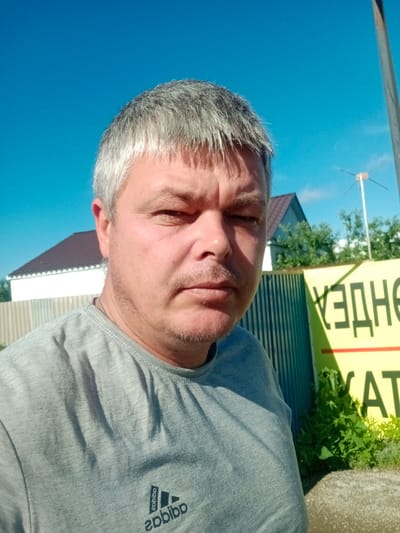 A middle-aged man with graying hair and a mustache is photographed outdoors on a sunny day. He is wearing a gray t-shirt with an Adidas logo. The background shows a blue sky, a house with a brown roof, a metal fence, green vegetation, and a yellow sign with black lettering, which appears to be in Russian. The sign contains the Cyrillic letters "ХЗДНЕ" and "АТ". There is also a radio antenna visible in the background. The man's expression is neutral, with his eyes partially closed. The image appears to have been taken in the city of Qobda, Kazakhstan, based on the presence of the Russian language on the sign, as Russian is widely spoken in Kazakhstan.