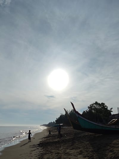 A bright, round sun is visible in a hazy sky above a sandy beach in Curehtunong, Indonesia. The sun is the brightest point in the image, partially obscuring the sky with its glare. The sea is calm with gentle waves lapping at the shore. On the beach, a lone child stands silhouetted against the water. Further down the beach, other figures, possibly children and adults, are present, though less clearly defined. Two fishing boats are pulled up onto the sand, one a vibrant teal and red, the other a duller yellow. Behind the boats, a line of trees forms a dark green backdrop against the sky. The overall impression is one of a serene, early morning or late afternoon scene.