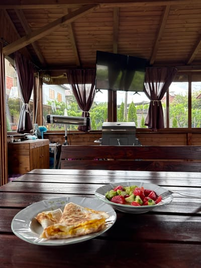 This media file captures a domestic scene within a wooden outdoor structure, likely a gazebo or enclosed patio, located in Polena, Bulgaria. The structure features wooden paneling on the ceiling and walls, with large windows offering a view of a residential area and lush greenery.
In the foreground, a dark, glossy wooden table is set with two plates of food. One light-colored, scallop-edged bowl contains a salad composed of diced red (likely tomato) and green (likely cucumber) ingredients, with a silver fork resting in it. Another matching plate holds several folded, light-brown food items, resembling crepes, omelets, or flatbreads.
To the left, a flat-screen television is mounted on a wall. Brown curtains are tied back at the windows, revealing the exterior. Outside, through the windows, a silver-colored gas grill is visible. Further in the background, a house with a tiled roof and light-colored walls can be seen amidst trees and other vegetation. A tall, silver and black electric heater stands on a base near the window. Below the heater, a blue brush and other cleaning supplies are placed on a wooden surface.
The scene depicts a peaceful mealtime setting during the daytime, indicated by the natural light, though the sky appears overcast or partly cloudy. No people are visible. Some faint, illegible text appears on a blue item near the cleaning supplies.
