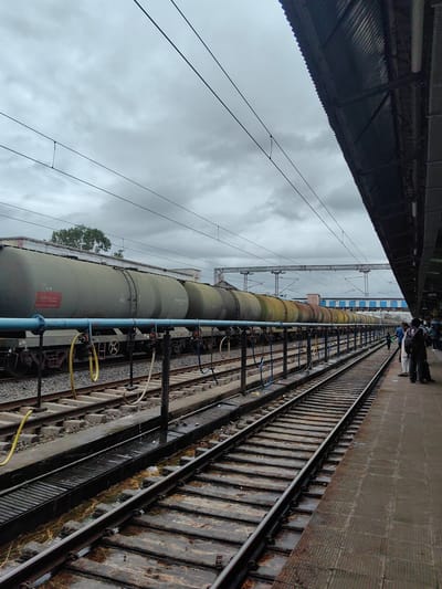 The image captures a cloudy day at the Itarsi railway station in India. The foreground shows multiple train tracks, with water pooled on some of the surrounding infrastructure, indicating recent rain or washing. To the left, a long train of olive-green tanker cars, labeled "PETROL" on one of the visible cars, is parked on a track adjacent to the platform. Blue and yellow hoses are connected to these tankers, suggesting the transfer of liquid cargo. The platform on the right has several people waiting, some with luggage. Overhead, power lines and the corrugated roof of the station shelter are visible against the overcast sky. The scene depicts a typical, albeit somewhat damp, operational railway environment.