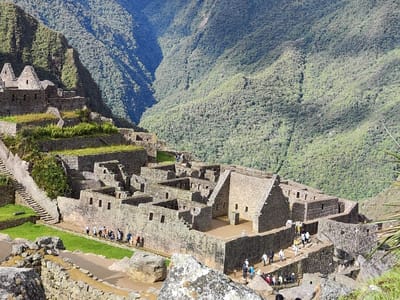 Machu Picchu, an ancient Inca citadel, nestles among lush green mountains. Stone ruins, featuring precise masonry, form buildings and terraced platforms. Tourists explore the site, walking along pathways and between structures. The ruins are set against a backdrop of a deep valley and densely forested slopes under a bright sky. The scene conveys a blend of human construction and the natural landscape.