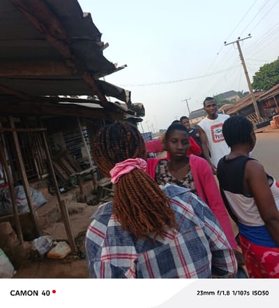 This street scene, set in Agbani, Nigeria, captures several individuals amidst rustic structures under an overcast sky, suggesting late afternoon or early morning. In the foreground, an individual with braided, reddish-brown hair tied with a pink scrunchie is seen from behind, wearing a blue and white plaid shirt. To their right, a young woman in a pink hooded jacket and a floral top looks down with a contemplative expression. Behind her, a man in a white t-shirt, emblazoned with the text "PRIDE," smiles slightly towards the viewer. Another person, partially visible on the far right, wears a dark tank top and a patterned skirt.
On the left side of the image, a makeshift stall or building, constructed from corrugated metal sheets and rough wooden planks, appears aged and somewhat dilapidated. Sacks and various items are scattered on the unpaved ground around the structure. The background features a dirt road stretching into the distance, flanked by utility poles with power lines. Simple, indistinct buildings and sparse vegetation line the road, indicating a semi-urban or rural environment. The overall scene depicts a casual moment of daily life on a local thoroughfare.
Visible text includes "CAMON 40" on the bottom left, likely indicating the camera model, and "23mm f/1.8 1/107s IS050" on the bottom right, detailing the camera's photographic settings.