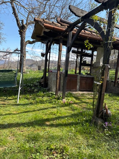 The image shows a partially constructed or rustic outdoor kitchen structure in a grassy yard. The structure features dark, heavy timber framing and a tiled roof. It has open sides with a window-like grid and a set of wooden cabinet doors set into a stone base. To the left, a bare tree branch extends into the frame, with a swing set hanging from it. In the background are distant hills or mountains under a blue sky. The yard is green with patches of flowers. A decorative wooden barrel sits in the grass next to the structure. The location is in Gorno Trape, Bulgaria.
