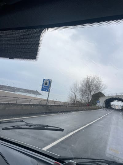 The image depicts a view from within a vehicle, looking forward through a rain-streaked windshield in Zumaia, Spain. A black windshield wiper rests horizontally in the lower-left section of the frame. The vehicle is on a wet, paved road with a continuous white line marking the right edge of the visible lane. To the left, a concrete barrier separates the road from a body of water, possibly the sea, with a distant breakwater visible. A blue rectangular road sign on a grey pole stands on the left side of the road, displaying a white tunnel pictogram and partial text, indicating a tunnel ahead. Further down the road, an arched tunnel entrance is visible. On the right side of the road, leafless trees and a building with a dark roof are situated prior to the tunnel entrance. The sky is overcast and uniformly grey, consistent with wet weather conditions. No individuals are clearly identifiable.