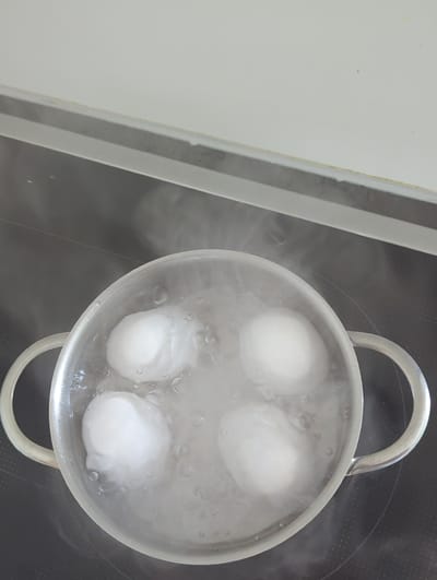 A close-up, overhead shot shows four white eggs in a pot of boiling water. The pot is silver with two handles and sits on a black stovetop. Steam rises from the water, obscuring some of the eggs. The stovetop has faint circular markings, suggesting it's an induction hob. To the left of the pot, a portion of a white countertop or cabinet is visible, with a thin grey and yellow stripe along its edge. The overall scene is clean and domestic, suggesting a kitchen setting. The lighting appears to be indoor, with no discernible indication of the time of day or weather conditions. The focus is solely on the cooking process. There is no visible text in the image.