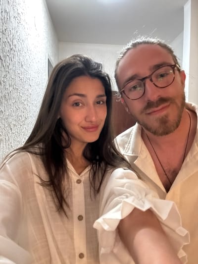 This is a close-up selfie-style portrait of two individuals, a man and a woman, positioned at a low angle looking upwards towards the camera. The image itself is rotated 90 degrees clockwise from its natural upright orientation.
The man, in the foreground on the left, has light brown hair, a beard, and a mustache. He wears distinctive round, brown-rimmed eyeglasses that reflect light. A thin, dark cord necklace is visible against his bare skin beneath his light-colored, collared shirt. The shirt, likely white or cream, features a subtle ruffled cuff detail on his sleeve.
Slightly behind him and to the right is the woman, who has long, dark brown hair that cascades over her shoulder. She is also wearing a light-colored, button-down shirt, with three brown buttons visible along the placket. Both subjects are looking directly at the camera with neutral to slightly smiling expressions, conveying a relaxed and pleasant mood.
The background consists of a textured, light-colored surface, possibly a wall or ceiling, indicating an indoor setting. The lighting is bright, with some backlighting apparent around the man's head. There is no visible text in the image. The scene suggests a casual moment captured between the two individuals. This media file is contextualized as being from Zankovići, Montenegro.
