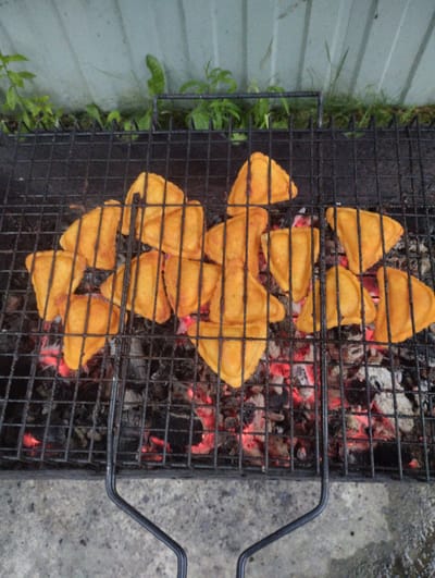 In Urkovo, Russia, a grill grate, positioned over glowing red embers and ash, holds a cluster of approximately fifteen golden-brown, triangular-shaped pastries. The pastries appear to be filled and are arranged haphazardly on the grate. Behind the grill, a light-colored corrugated metal fence is visible, with some green foliage growing in front of it. The ground beneath the grill is a rough, concrete surface. The scene suggests an outdoor cooking environment.