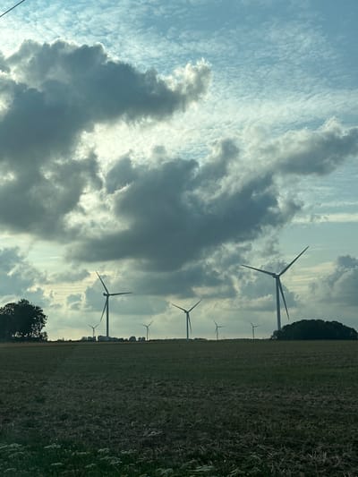 This image depicts a rural landscape with multiple wind turbines silhouetted against a cloudy sky. The foreground is a dark, grassy field, possibly agricultural. In the mid-ground, a line of wind turbines stands on a slightly elevated horizon, interspersed with some trees and what appear to be buildings or structures. A larger cluster of trees is visible on the left side of the frame. The sky is a mix of bright blue patches and heavy, gray clouds, with sunlight breaking through in some areas. The overall impression is of a windswept, open environment. This image was taken in the vicinity of Saint-Maclou-de-Folleville, France.
