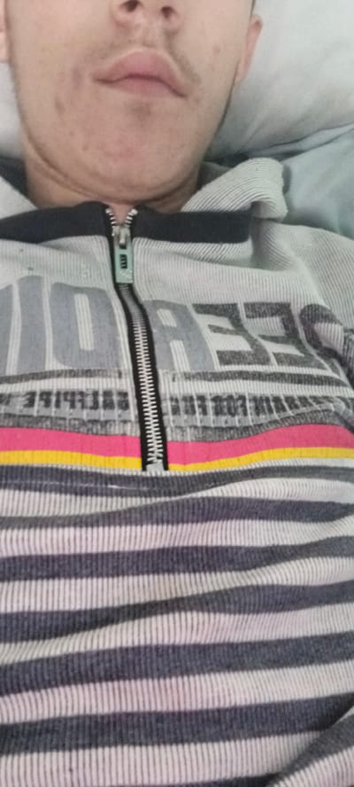 The image shows a close-up view of a person's chin, mouth, and chest, wearing a grey and white horizontally striped hoodie with a zipper that is partially open. The hoodie has text and graphics on the front, including the words "IDOL" and "DEES" in a distressed font. There are also colorful horizontal stripes in pink and yellow above a section of horizontal stripes on the hoodie. The person has some facial hair and what appears to be light acne on their chin. The background is out of focus and appears to be a light-colored pillow or bedding. There is no discernible information about the location in the city of Булум, Russia.
